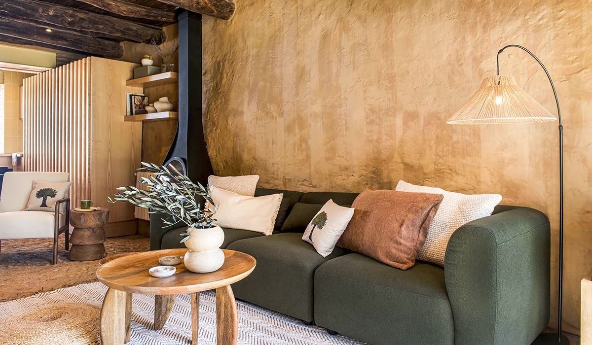 Sofá verde con cojines neutros, mesa de madera y lámpara de fibras junto a pared de barro en vivienda de la Sierra de Aracena